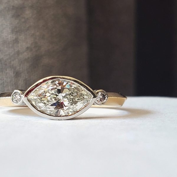 A marquise diamond bezel set east west in platinum with two bezel set round brilliant diamonds on a 14k yellow gold knife edge band with millgrain detail.