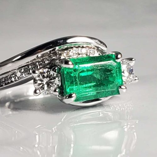 Three stone bypass style 14k white gold engagement ring with a Columbian Emerald, diamond sides and diamond accented twisted band, and a thin half-round wedding band. Created by Katherine Spencer. Made in Morgantown, West Virginia USA