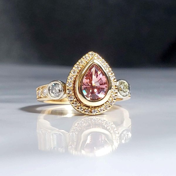 Stunning pear shaped champagne pink garnet, bezel set in a diamond halo with two round bezel set diamonds on either side, all set in 14k yellow gold.