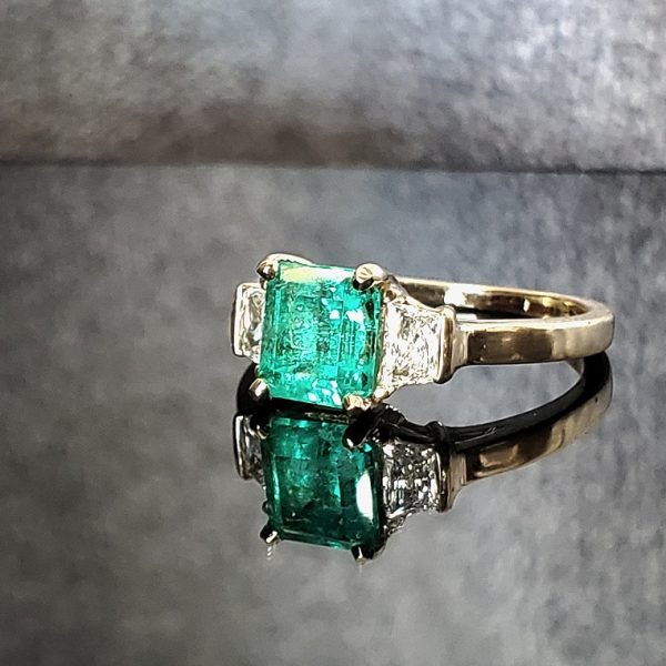 A large center emerald prong set in 18k yellow gold with trapezoid diamonds tapering down to a delicate flat band