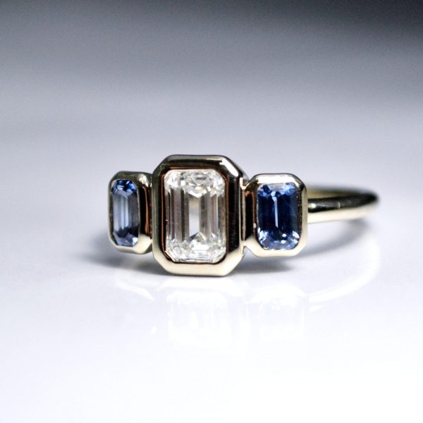 Stunning 1.07 ct. emerald cut diamond accompanied by two light blue emerald cut sapphires all bezel set in platinum.