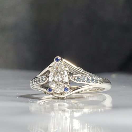 Cathedral style engagement ring with a pear shape diamond, blue sapphire accents in the prongs and blue aquamarine accent gemstones, set in 14k white gold. Created by Katherine Spencer. Made in Morgantown, West Virginia USA
